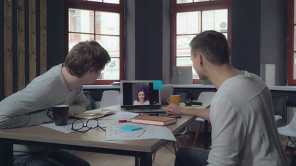 Two businessman in the modern office startup company have video conference call with colleague or friend of clent. Men using application looking on the screen laptop talking with woman. Casual