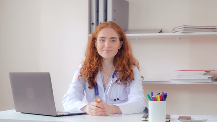 Obraz na płótnie Canvas Portrait redhead doctor sitting at the desk in office. Candid professional woman therapist posing looking at the camera and friendly smiling.