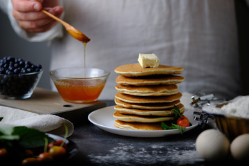 Man pours honey delicious pancakes. On the table are the products from which they prepared the dish and berries. Dad prepares a delicious breakfast. The hands of the cook in the frame.
