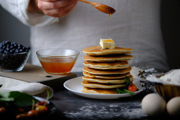 Man pours honey delicious pancakes. On the table are the products from which they prepared the dish and berries. Dad prepares a delicious breakfast. The hands of the cook in the frame.