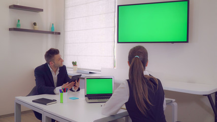 Adult handsome worker wearing elegant suit using digital tablet showing presentation or new design project. On the desk laptop with green screen. Back view on the businesswoman