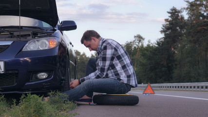 Caucasian man changing a tire sitting on stepney near car on the roadside. Handsome traveller outdoors