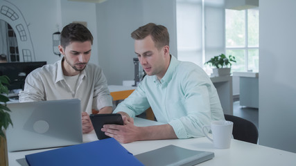 Concentrated contemplative man at the working place using digital tablet. His partner young professional worker search information in computer. Coworkers working in modern creative office.