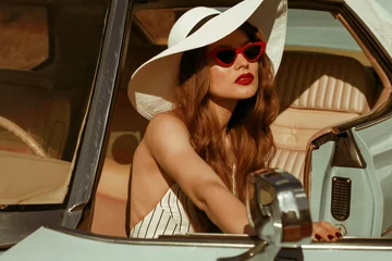 Fotobehang Street fashion portrait of young elegant luxury lady wearing white sunglasses, wide brim hat, striped linen jumpsuit, posing in the retro car. Copy, empty space for text © Victoria Fox