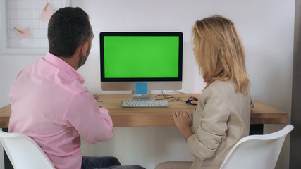 Business people sitting at the desk in front of computer with green screen have conversation. Meeting in the office with white wall and casual interior
