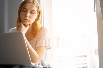 young girl sitting near the window with a laptop, she uses the computer at home on the windowsill, a woman freelance, copy space for text