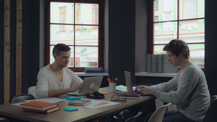 Two young businessmen working in the modern contemporary office with big windows. Group of people works with computer sitting at the wooden desk with paper documents. Happy startup team.