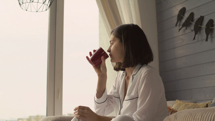 Portrait woman sitting near the big window holding red cup. Lady wearing in white pajamas sits in...