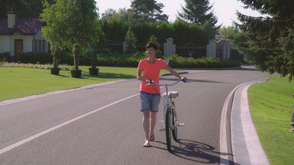 Caucasian woman with bike in the city. Happy adult brunette wearing shorts and t-shirt enjoy active leisure at summertime. Lady looking nature view.