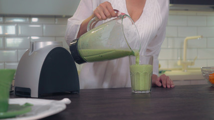 unrecognizable woman pouring green vegetables juice in glass. Female hand holding blender with detox smoothie at home.