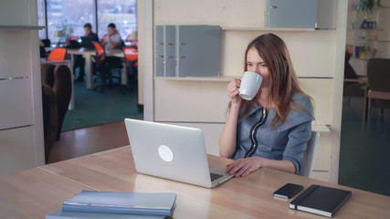 Obraz premium Woman typing on the laptop drink coffee. Female smiling and happy about her project. She wearing in elegant formal business dress.On the background work two men colleagues