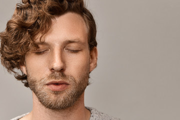 Obraz premium Close up portrait of handsome unshaven young male with stylish hairdo and freckles having peaceful calm facial expression keeping eyes closed, practicing meditation in the morning. Harmony and balance