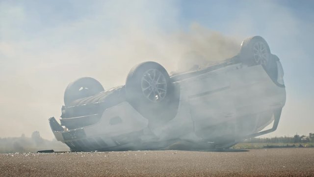 Horrific Traffic Accident Rollover Smoking and Burning Vehicle Lying on its Roof in the Middle of the Road after Collision. Daytime Crash Scene with Damaged Car. Low Angle Sunny Day Shot
