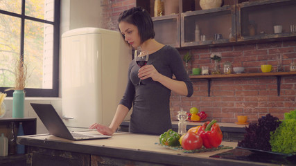 Happy young woman drinking red wine at home. Lady standing near the big window in the kitchen using laptop surfing internet. Businesswoman looking on the screen read or order delivery food she could