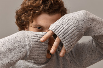 Human facial expressions, emotions and body language. Mysterious guy with curly ginger hair wearing cozy long sleeved gray sweater keeping hands in front of him as if hiding, staring at camera