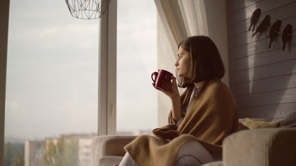 Woman sitting on the chair near the big window. Young lady wearing in white pajamas wrapping up in blanket drinking tea or coffee in the morning at home. Caucasian model looking on the street and