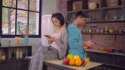 Young couple using gadgets in kitchen at home. Caucasian woman texting message on smartphone. Man holding touch screen tablet surfing internet or check mail wearing in hoodie. Relationships in family