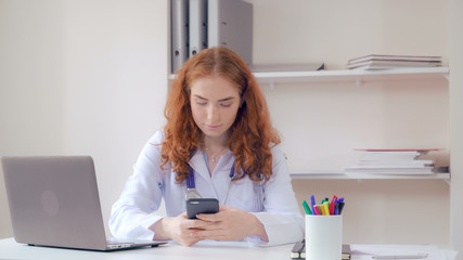 Redheaded doctor sitting in the office in hospital. Woman holding smartphone messaging. Therapist using app on mobile phone typing message. Professional nurse chatting in clinic.