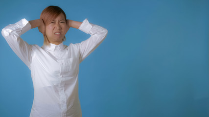 Naklejka premium portrait young asian female hears a terrible sound or scream on blue background in studio. attractive korean woman with blond hair wearing white casual shirt looking at the camera.