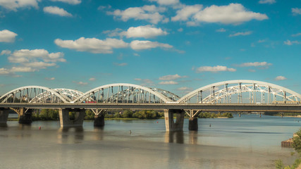 The movement of clouds over the bridge along which cars and trains are moving, a river flows under...