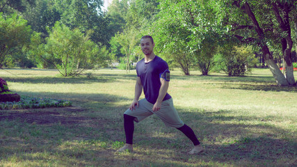 A handsome man in trendy outfit is sitting down on his right leg to warm up before the training. He is wearing trendy shorts and leggins outfit paired with a blue T-shirt and an armband for the