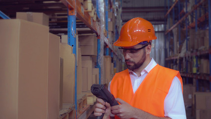 Worker wearing high visibility clothing and a hard hat scans labels on boxes using wireless barcode scanner. Handsome man working at warehouse.
