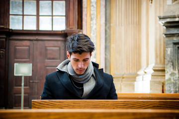 Young man sitting and kneeling in church praying, on wood bench
