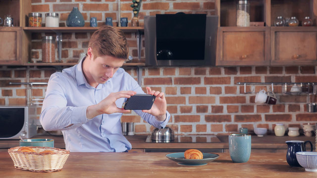 Handsome caucasian businessman taking photo his breakfast. Smiling man using smart phone using application shared picture in social media. Happy man wearing elegant suit in studio apartment. Morning