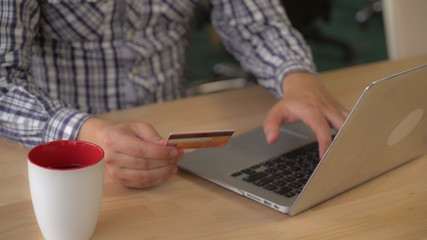 man hands buy online via internet on laptop. Guy sitting at the wooden desk with pc and mug with tea or coffee in the office. Male holding credit card enter security code pay or order some purchase