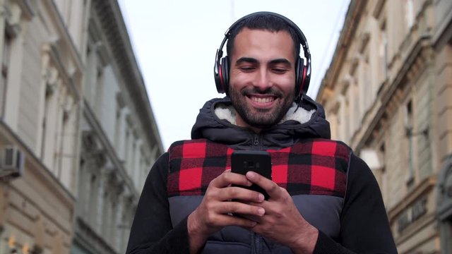 Multinational bearded Man is dancing on the European city&rsquo;s street in Headphones. He&rsquo;s using Musical Apps on his Smartphone. He&rsquo;s enjoying Music and smiles. Happy Emotions. Social Network. Music.