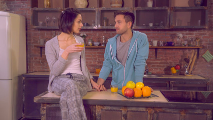 Beautiful couple in love enjoy morning in apartment. Young family drinking orange juice on breakfast. Attractive lady speaking with handsome man in kitchen. Two people talking discussing plans for day