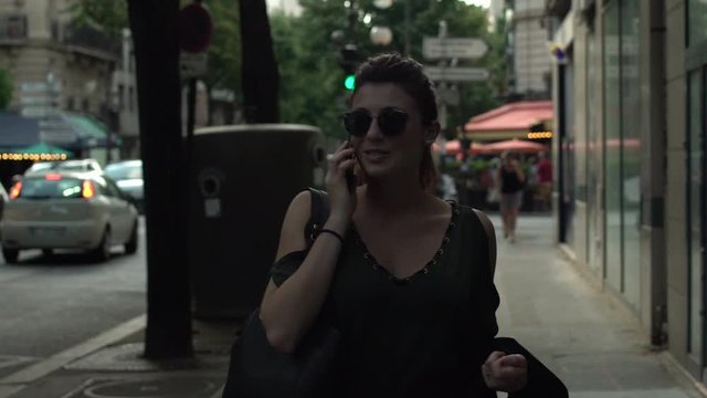 Attractive caucasian woman with sunglasses, freckles, piercings and red hair having phone conversation walking through the street, during sunny summer in Paris. Slow motion. Stylish girl.