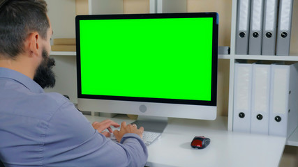 businessman sitting at workplace in small office. man with beard looking on display computer with green screen. employee in startup company typing on keyboard - Powered by Adobe