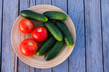 Fresh vegetables in a wooden, round plate. Red tomatoes and green cucumbers in a bamboo bowl. on the background of the wooden table of their old boards. Summer evening.