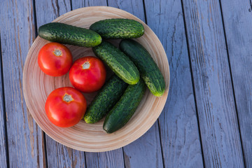 Fresh vegetables in a wooden, round plate. Red tomatoes and green cucumbers in a bamboo bowl. on the background of the wooden table of their old boards. Summer evening.