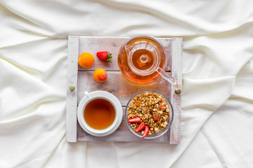 Breakfast in bed with granola, tea in pot and cup on tray on white bed background top view