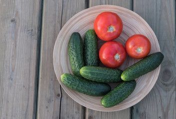 Fresh vegetables in a wooden, round plate. Red tomatoes and green cucumbers in a bamboo bowl. on the background of the wooden table of their old boards. Summer evening.