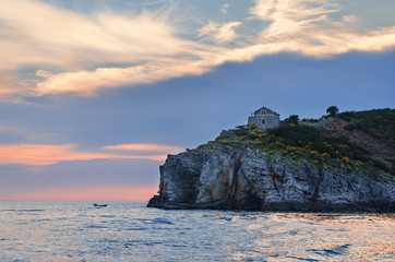 Lonely little house on a stone precipice over the sea against the backdrop of sunset. A small fishing boat in the open sea. A beautiful seascape on the Adriatic.