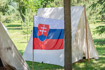 Slovakia flag and camp tent in a forest in nature on a sunny summer day. Camping and retro tent under shadow of pine forest in Europe with state symbols. Close up, selective focus