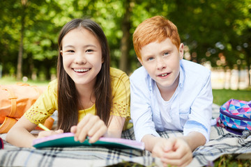Two smiling kids lying on grass in park, having fun while doing homework together after classes
