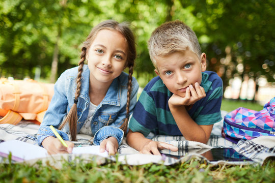 Two Smiling Kids Chilling After School On Grass In Park While Doing Homework Together Outside