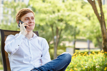 Young attractive man in white shirt sitting on brown chair in the park on green background - 277463929