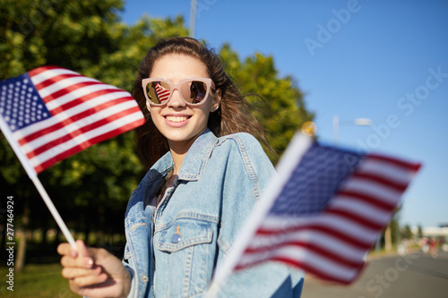 Portrait of jovial student girl in pink sunglasses and denim jacket walking over street and holding national flags