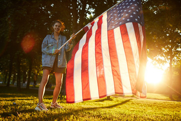 Content confident pretty girl in long denim jacket standing in forest and waving national flag at sunset