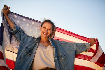 Portrait of happy excited young woman in denim jacket holding US flag against blue sky
