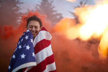 Cheerful excited young woman wrapped in American flag standing against firework and keeping eye closed to protect them from colored smoke