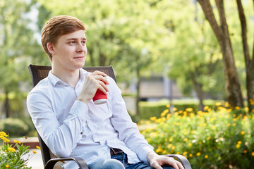 Young attractive man in white shirt sitting on brown chair in the park on green background - 277466343
