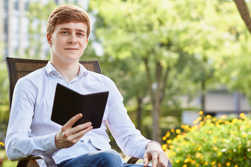 Young attractive man in white shirt sitting on brown chair in the park on green background - 277466912