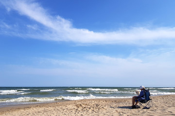 Man with hat sitting on a beach chair and holding a beer bottle, on a beach next to a sea
