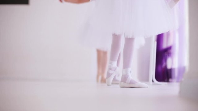 Two young ballerinas repeat after the teacher exercises for legs near barre stand, legs closeup. Children girls are in white tutus and pointes stand on tiptoe training ballet elements.